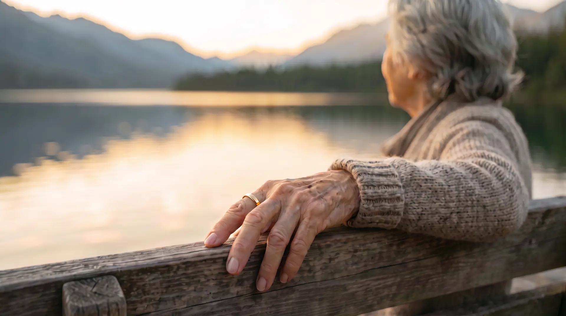 An older woman sitting on a bench by a mountain lake, with a gold ring on her index finger